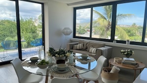 Un espace repas moderne avec une table en verre, des chaises blanches et une vue sur la verdure environnante.