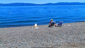 Una playa pedregosa con una persona en silla de ruedas y otra sentada en una silla de playa, un perro, y un cielo azul despejado con montañas a lo lejos.