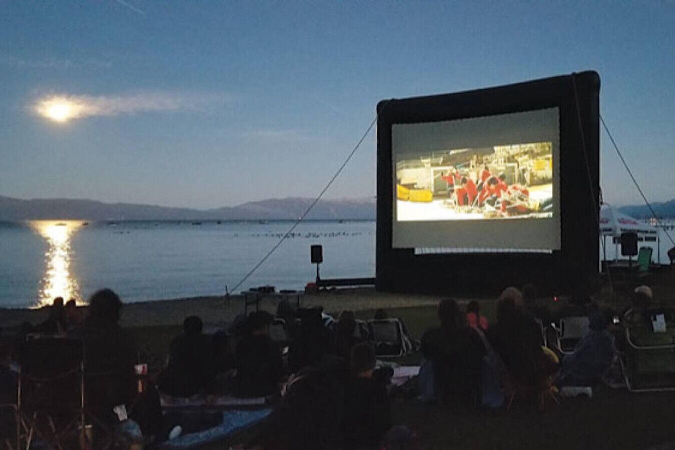 Eine Freiluftleinwand mit einer Gruppe von Zuschauern davor, mit Blick auf ein Gewässer.