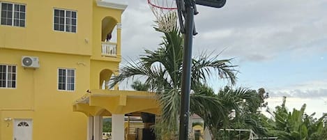 A basketball hoop in front of a yellow building with a covered seating area and a swing set.