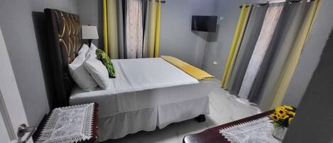 A small, neatly arranged bedroom with a bed, bedside tables, a TV, and curtains.