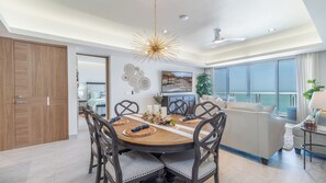A modern dining area with a round table, dark chairs, and a chandelier. There is a balcony with a view of the sea.