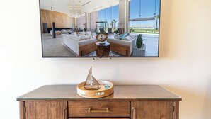 A modern living room with a large TV mounted on the wall, a wooden cabinet with a decorative item, and a view of the ocean through large windows.