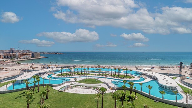 A resort with a large central pool, palm trees, and a view of the ocean.