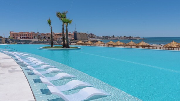 A large, clear swimming pool with a palm tree and thatched umbrellas, surrounded by buildings and a beach.