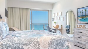 A hotel room with a large bed, a desk, a chair, a dresser, and a view of the ocean.