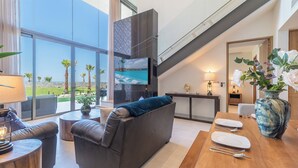 Un salon moderne doté d’une grande fenêtre, d’une cheminée, d’un coin repas avec une table dressée pour deux personnes, et offrant une vue sur une piscine et des palmiers.