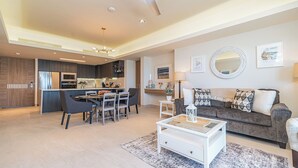 A modern living room with a grey sofa, white coffee table, and a dining area with chairs and a kitchen in the background. - One 1607 - Premier Beach Destination Blending Comfort with Luxury (Puerto Peñasco)