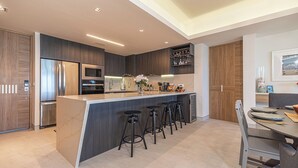 A modern kitchen with a central island, bar stools, and a dining area with a table and chairs. - One 1607 - Premier Beach Destination Blending Comfort with Luxury (Puerto Peñasco)