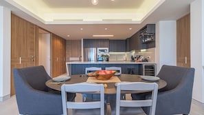 A modern kitchen with a central island, built-in appliances, and a dining area with grey chairs and a wooden bowl of fruit.
