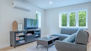 A modern living room with a grey sofa, a coffee table, and a TV on a stand. There is a bookshelf with books and a decorative basket. - Sauna Retreat Near Burk's Falls | Games Room • Bocce Court • Fire Pit • Starlink (Burk's Falls)