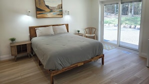 A bedroom with a bed, a nightstand, a chair, a painting, and a view of the outdoors. - Sauna Retreat Near Burk's Falls | Games Room • Bocce Court • Fire Pit • Starlink (Burk's Falls)