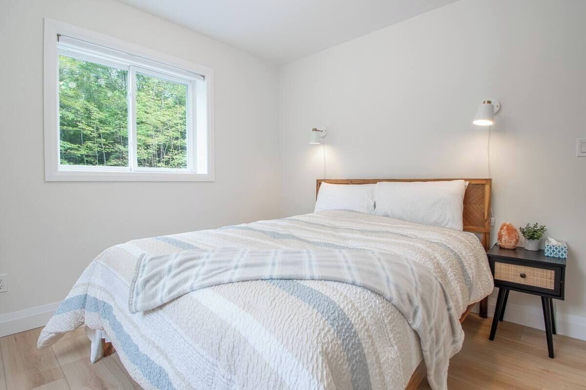 Una camera da letto con un letto, lampade da comodino, un tavolino e una finestra vista alberi, con un interruttore a muro.