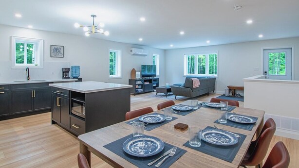 A modern kitchen with dark cabinets, a central island, and a dining area with a wooden table set for four. - Sauna Retreat Near Burk's Falls | Games Room • Bocce Court • Fire Pit • Starlink (Burk's Falls)
