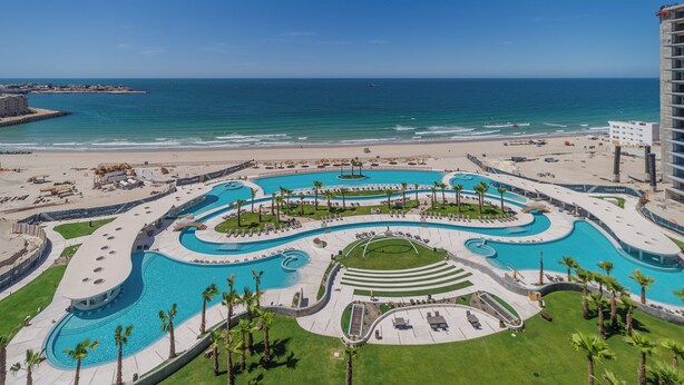 Een resort met een groot, kronkelend zwembad, omgeven door palmbomen en een strand.