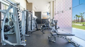 A modern gym with various exercise equipment, a wall-mounted TV, and a view of a landscaped area with palm trees. - One 1002: Hotel Room– Luxury Oceanfront Condo with Stunning Views (Puerto Peñasco)