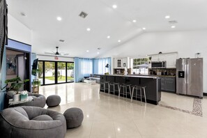 A modern kitchen with a bar area, a dining space with grey furniture, and a view through the glass door. - Luxury Oasis for 11: Pool & Games, 15 Min to Beach (Fort Lauderdale)