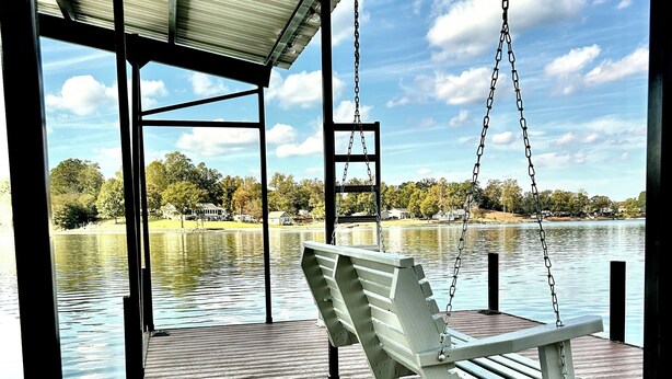 A wooden deck with a swing chair overlooking a calm lake, surrounded by trees and houses.