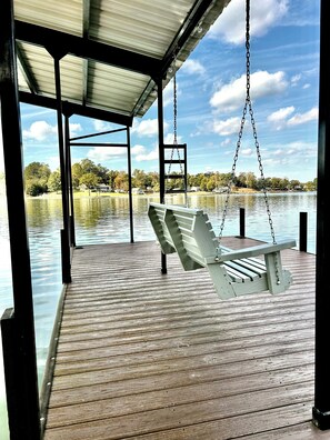 A wooden deck with a swing chair overlooking a calm lake, surrounded by trees and houses.