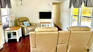 A room with a beige leather sofa, a small wooden table with a lamp, a flat-screen TV mounted on the wall, and large windows with curtains.