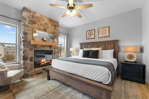 A bedroom with a stone fireplace, a bed with a quilted mattress, a nightstand with a lamp, and a ceiling fan. - Table Rock View Log Cabin with Private Hot Tub, Firepit, Lake Access & Free Attraction Tickets (Branson)