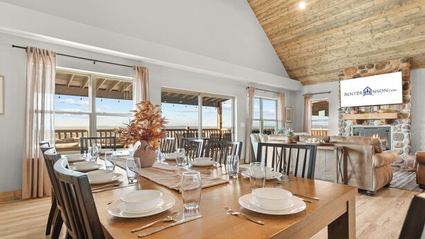Un coin repas avec une table en bois dressée pour quatre personnes, de grandes fenêtres et une cheminée en pierre.