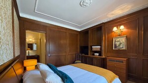 Quarto de hotel com uma cama grande, painéis de madeira, televisão, estante de livros e uma pintura na parede.
