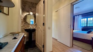 Casa de banho com lavatório, espelho e porta que dá para um quarto com cama e janela.