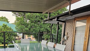 Een buitenpatio met een tafel met glazen blad en witte stoelen, omringd door een houten terras en een overdekt gedeelte met een bakstenen muur en een glazen deur.