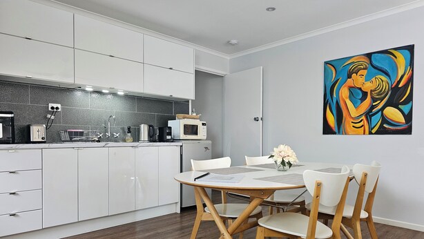 Een moderne keuken met witte kastjes, een magnetron en een koffiezetapparaat. Er staat een eettafel met stoelen en er hangt een schilderij aan de muur.