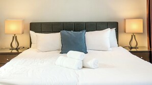 Un lit bien fait, avec du linge de lit blanc, une tête de lit rembourrée de couleur grise et deux lampes de chevet.