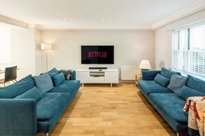 A modern living room with two blue sofas, a white entertainment unit, and a large flat-screen TV displaying 'NETFLIX'. - Spacious 2 Bed Flat with Garden for up to 6 (Greater London)
