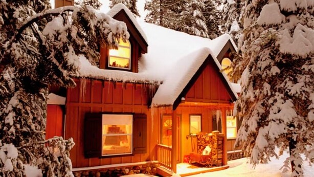 Une cabane en bois dont les fenêtres diffusent une lumière chaleureuse, entourée d’arbres couverts de neige.