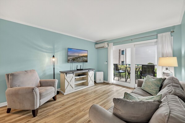 Ein Wohnzimmer mit beigefarbtem Sessel, grauem Sofa, Holzboden, Fernseher auf einem weißen Schrank und einem Balkon mit Gartenmöbeln.