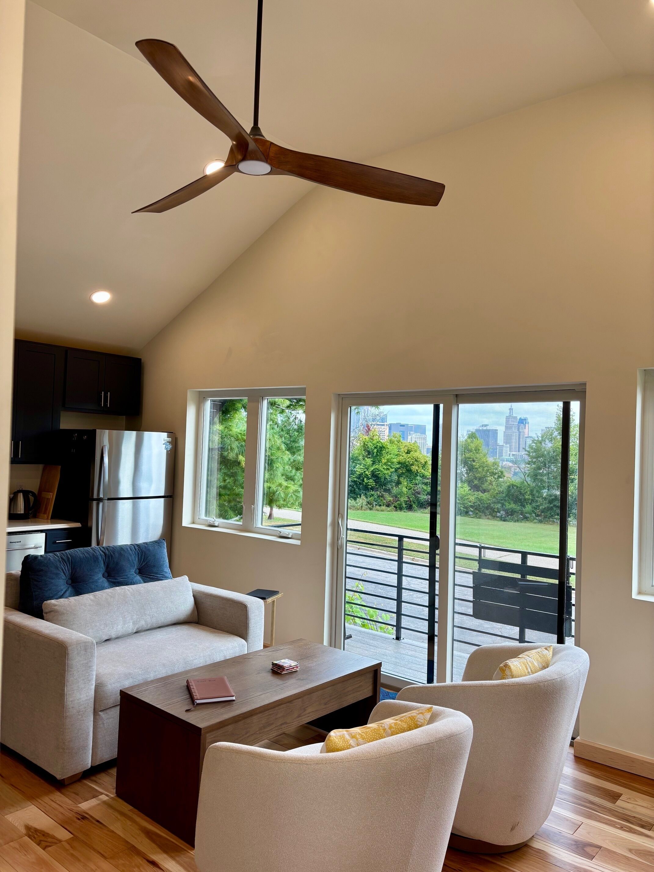 Un salon spacieux doté d’un ventilateur de plafond, d’un canapé, d’une table basse et offrant une vue sur le paysage urbain grâce à de grandes fenêtres.