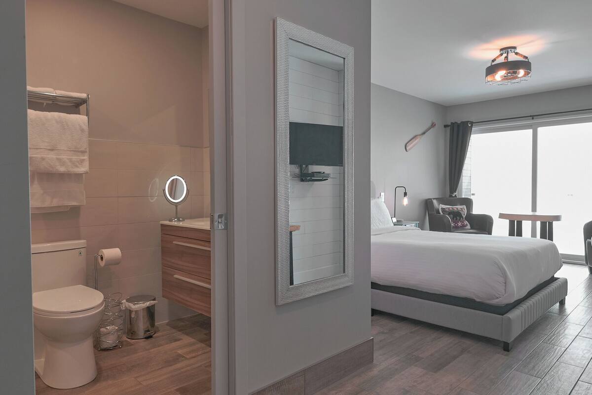 A modern bedroom with a large bed, a bedside table, a mirror, and a window with curtains.