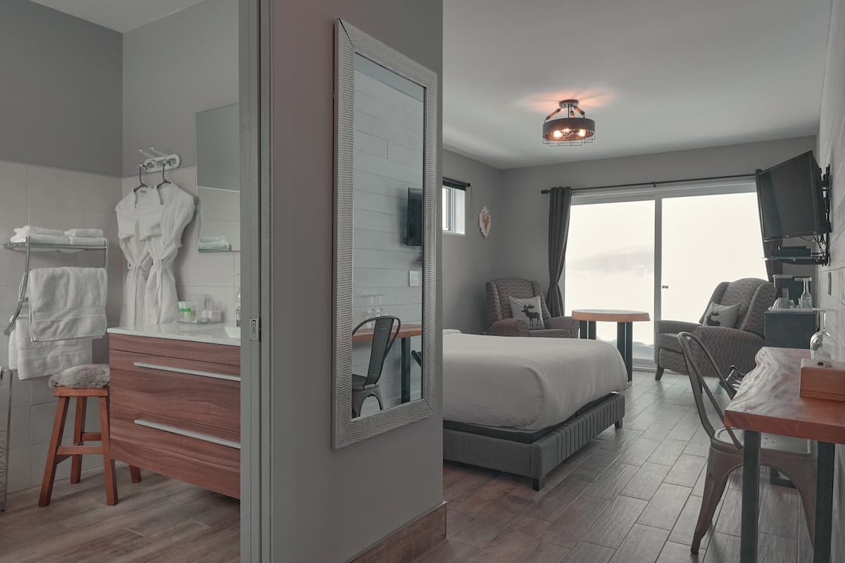 A modern hotel room with a large bed, a vanity with a mirror, a small table, a chair, and a view of the outdoors through a large window.