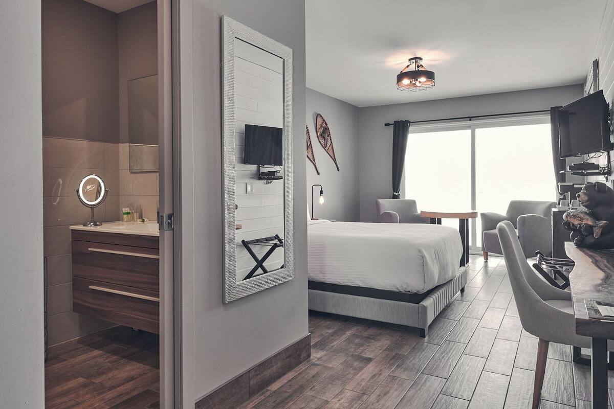 A modern hotel room with a large bed, a desk with a chair, a TV, and a bathroom with a mirror and vanity.