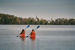 Two people kayaking on a calm lake with houses and a church in the background. - L'étoile du lac Lyster (Coaticook)