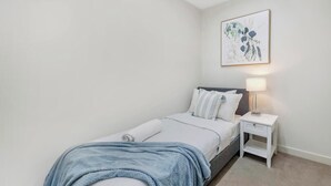 Ein Einzelbett mit blauer Decke, ein Nachttisch mit Lampe sowie ein gerahmtes Blumenbild an der Wand.