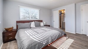 Une chambre à coucher comprenant un lit, des tables de chevet, une fenêtre avec des stores, une porte et une vue sur une salle de bain.