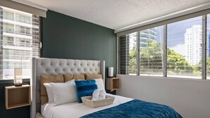 A bedroom with a tufted headboard, a large window with a view of buildings and trees, a bedside table with a lamp, and a basket of towels.