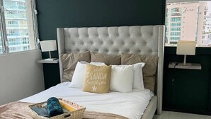 Een net opgemaakt bed met een geborduurde hoofdbord, een nachtkastje met een lamp en een raam met uitzicht op gebouwen.