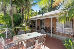 A covered patio with a table and chairs, surrounded by lush greenery and a brick building.