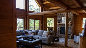 A cozy cabin interior with a stone fireplace, wooden beams, and a large window overlooking the outdoors. - Splash Mountain 6 bedrooms with indoor pool & mountain and filtered lake view (Garrett County)