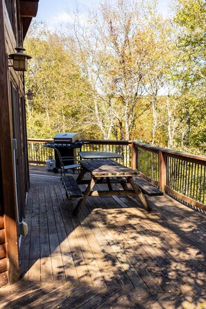 Une terrasse en bois avec un barbecue et une table de pique-nique, entourée d’arbres.