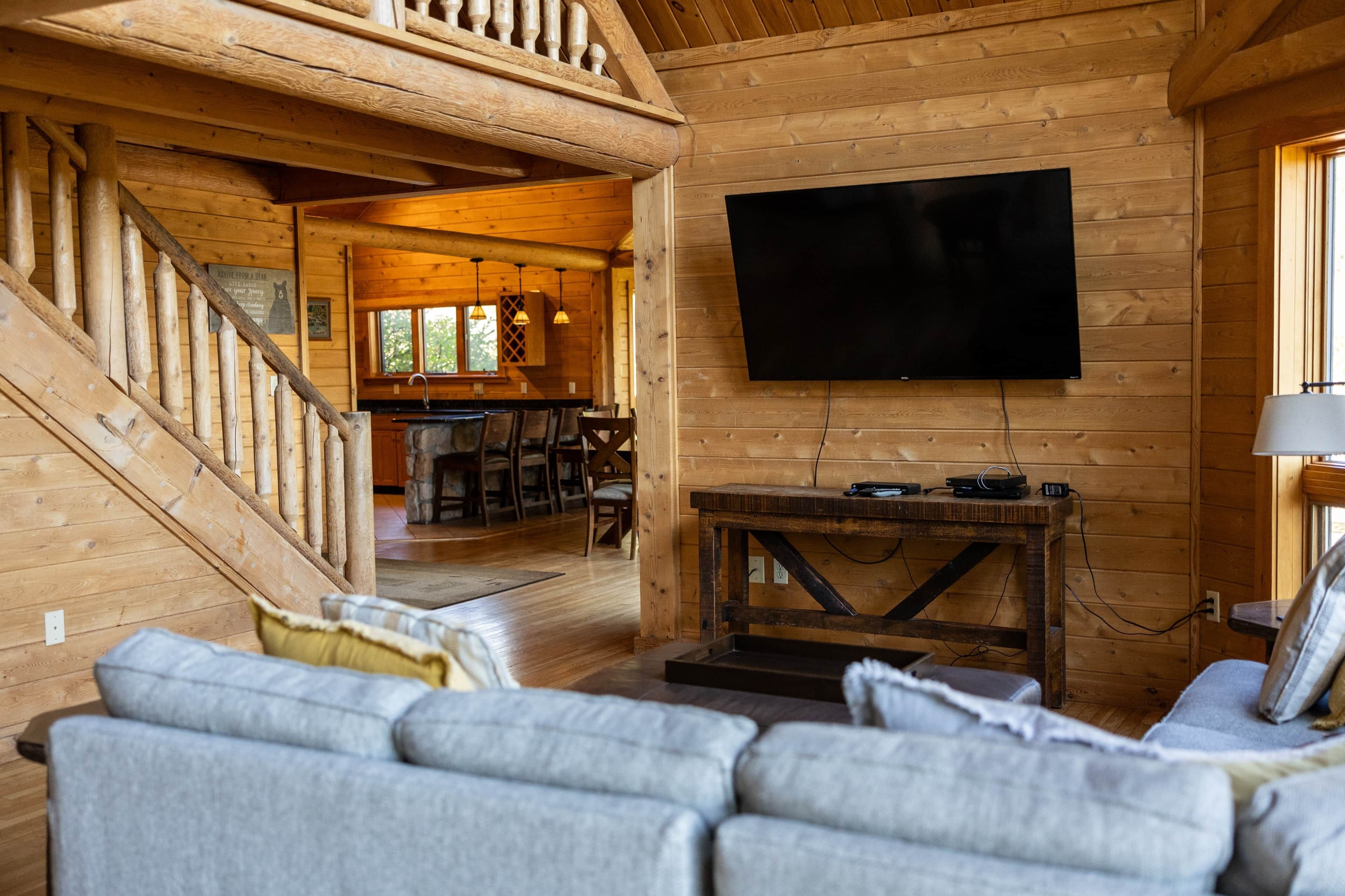 A wooden cabin interior with a living room, a staircase, a kitchen, and a TV mounted on the wall.