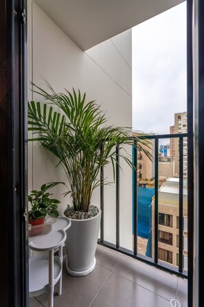 A balcony with two potted plants, a small table, and a view of buildings. - Geko | Bright 1BR near Larcomar w/ Pool and Gym (Miraflores)
