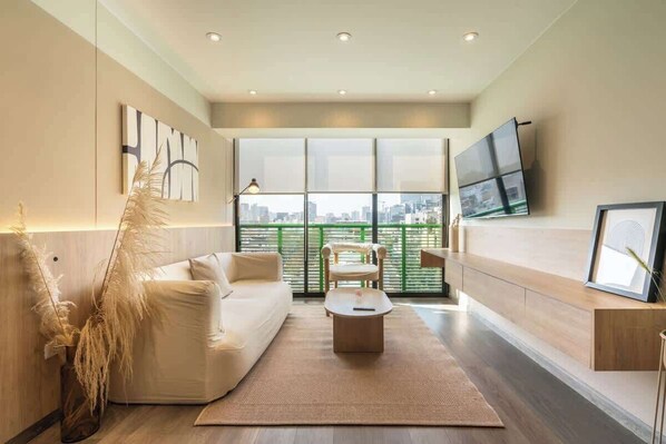 Ein modernes Wohnzimmer mit einer Couch, einem Couchtisch, einem Sessel, einem an der Wand montierten Fernseher und einem großen Fenster mit Blick auf die Stadt.