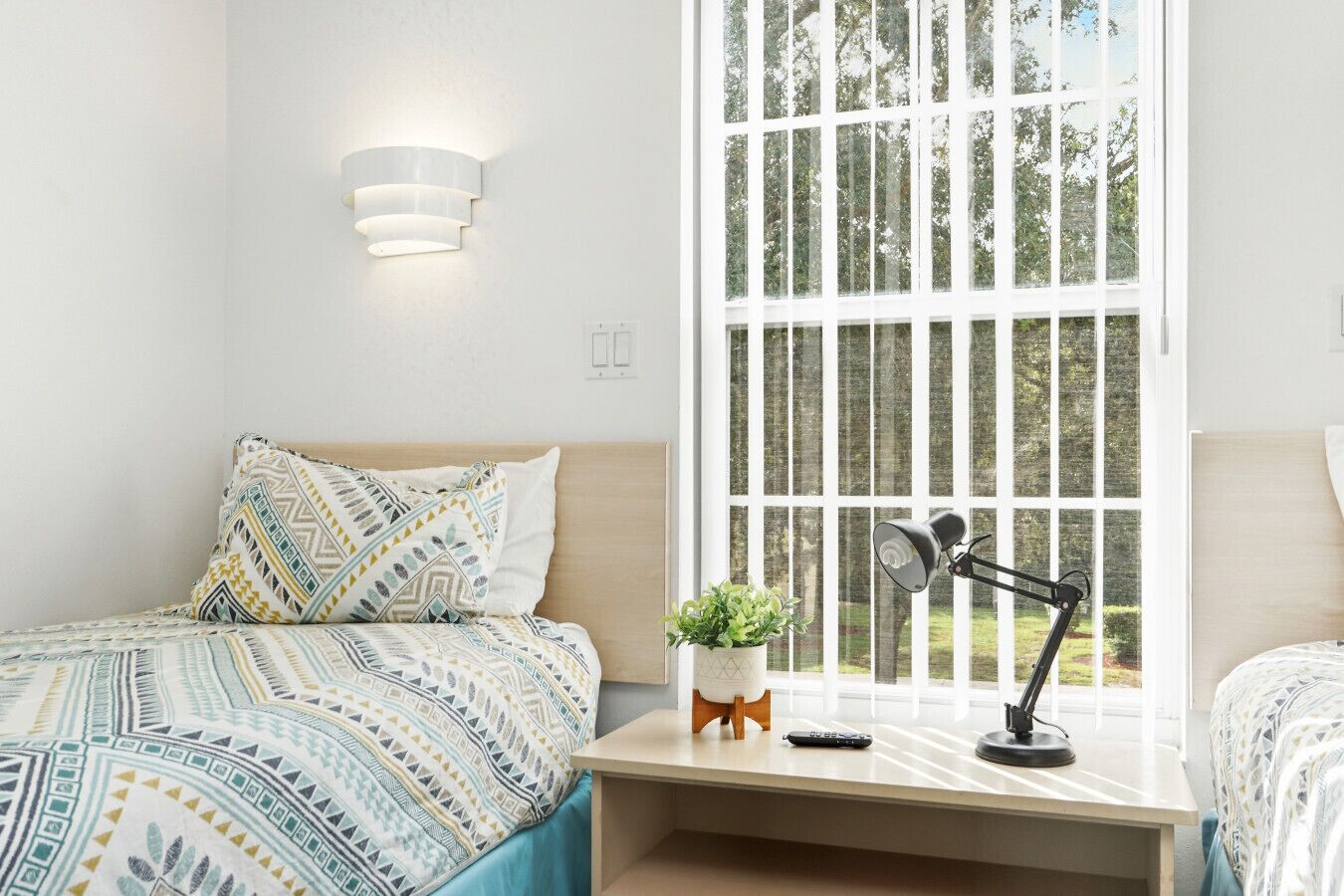 Une chambre avec une grande fenêtre, une table de chevet avec une lampe, une télécommande et une plante en pot.